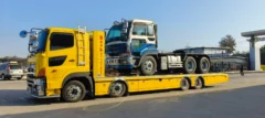 Yellow tractor-trailer with a blue-and-white semi on a flatbed trailer in an industrial yard under a clear blue sky.